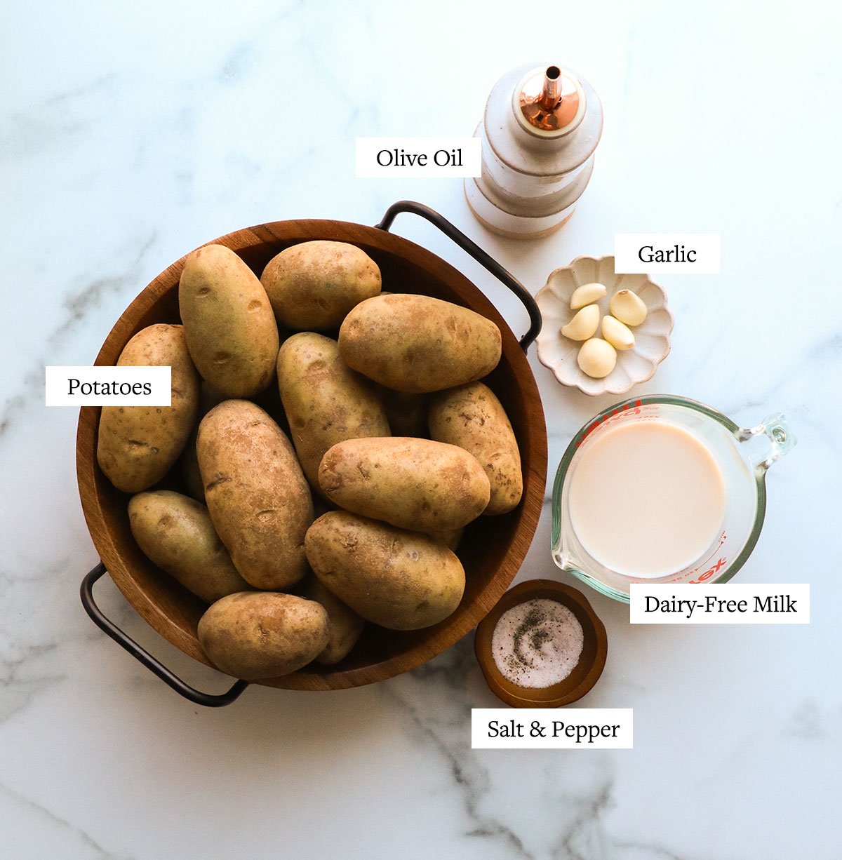 russet potatoes in a bowl near a cup of non-dairy milk, olive oil, garlic cloves, and spices.